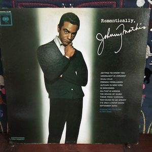 May include: Vintage vinyl record album cover featuring Johnny Mathis in a black and white photo. The album is titled "Romantically, Johnny Mathis" with a list of songs on the back. The cover has a dark green background.