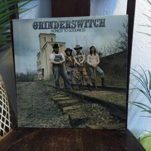 May include: Album cover for Grinderswitch's "Honest to Goodness." The image features four men standing on railroad tracks in front of a weathered building. The album title and band name are printed at the top.