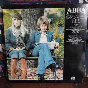 May include: ABBA Greatest Hits album cover featuring two people seated on a bench. The person on the left wears a denim jacket, jeans, and brown boots. The person on the right wears a denim jacket and jeans. The album cover is black with white text.