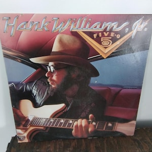 May include: Album cover for Hank Williams Jr. Five-O. The album art features a man wearing a hat and glasses, playing a guitar inside a car. The album title is in silver lettering, with a gold and brown graphic.