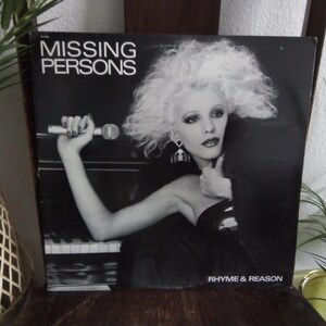 May include: Black and white album cover for Missing Persons, featuring a woman with voluminous white hair holding a microphone. The album title is at the top, and the album name "Rhyme & Reason" is at the bottom.
