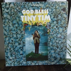 May include: Vintage vinyl record album cover for "God Bless Tiny Tim." The album art features a portrait of Tiny Tim holding a ukulele, set against a sky and floral background. The album cover has a blue and white marbled pattern.