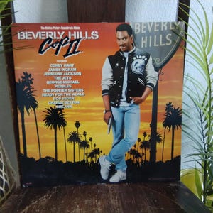 May include: Vintage "Beverly Hills Cop II" soundtrack album cover. Features a man in a letterman jacket and jeans, standing in front of a sunset scene with palm trees. The album title and artist names are prominently displayed.