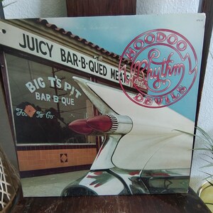 May include: Vintage vinyl record album cover featuring a retro car tail fin and a restaurant facade. The album title is "Hoodoo Rhythm Devils" in a pink circular design. The restaurant sign reads "Juicy Bar-B-Qued Meats" and "Big T's Pit Bar B Que".