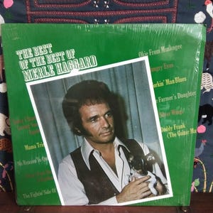 May include: A vintage vinyl record album titled "The Best of Merle Haggard." The album cover is green with a photo of Merle Haggard holding a small dog. The album's track list is visible on the right side of the cover.