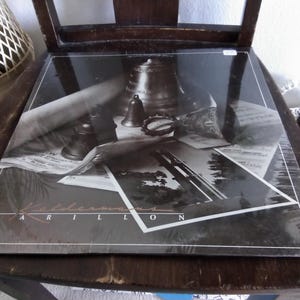 May include: A vinyl record album with a black and white cover featuring a still life of bells, sheet music, and a photograph. The album is titled "Carillon" with the artist's name "Keldermans" visible on the cover. The album is resting on a wooden chair.