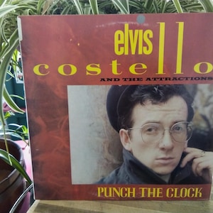 May include: Album cover for "Punch the Clock" by Elvis Costello and The Attractions. The cover features a portrait of Elvis Costello wearing glasses and a hat, set against a red background. The album title and artist name are in yellow.