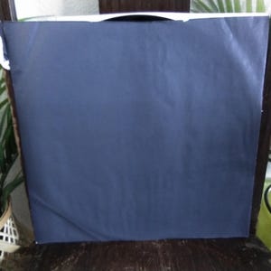 May include: A navy blue, rectangular object, possibly a package or artwork, is placed on a wooden chair. The object is wrapped in a smooth, dark blue material. Green plants are visible on either side of the chair.