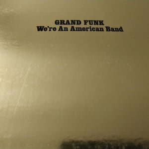 May include: A gold-colored album cover with the text "GRAND FUNK" above "We're An American Band" in black lettering. The background has a subtle, reflective sheen.