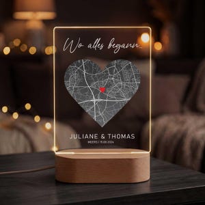Könnte beinhalten: Ein personalisiertes LED-Nachtlicht mit Holzsockel. Die transparente Acrylplatte zeigt eine herzförmige Karte mit roter Markierung, den Text "Wo alles begann..." und die Namen "JULIANE & THOMAS" mit einem Datum. Das Licht ist an.