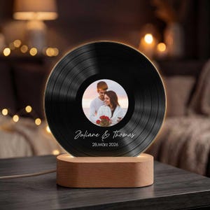 Può includere: Un disco in vinile nero con una foto di una coppia al centro, illuminato da una luce calda. Il disco è montato su una base di legno. I nomi "Juliane & Thomas" e la data "28.März 2026" sono stampati sul disco.