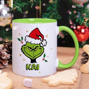 Könnte beinhalten: Weißer Keramikbecher mit grünem Innenraum und Griff. Der Becher zeigt ein grünes Grinch-Gesicht mit Weihnachtsmütze und dem Namen "KAI" darunter. Stechpalmen- und Sternakzente vervollständigen das Design, perfekt für Weihnachten.