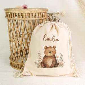 Op de afbeelding: Een beige trekkoordtas met een bruine berenillustratie en de naam "Emilia" in bruin schrift. De beer is omgeven door bloemen en planten. Een rieten mand staat op de achtergrond.