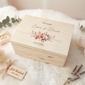 Puede incluir: Una caja de madera de color claro con un diseño floral, los nombres "Emma & David" y la fecha "05.07.2026" impresos en la tapa. La caja también tiene las palabras "FÜR IMMER" impresas en la tapa. Una etiqueta "Just Married" está atada a la caja con una cinta.