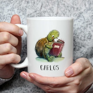 Può includere: Tazza in ceramica bianca con una tartaruga dei cartoni animati che legge un libro. La tartaruga è verde e marrone e il libro ha una copertina rossa. La tazza ha il nome "CARLOS" stampato in nero.