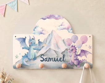 May include: A wooden wall-mounted coat rack featuring a watercolor illustration of a blue dragon and a purple dragon with balloons in front of mountains. The name Samuel is printed in dark blue script in the center. Four round wooden pegs are attached.