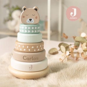 Peut inclure: Un jouet d'empilage en bois avec une tête d'ours et cinq anneaux colorés. Les anneaux sont dans des tons de beige, vert menthe et blanc, avec le nom "Carlos" gravé sur l'un d'eux. Le jouet est conçu pour les enfants.