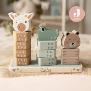Può includere: Giocattolo impilabile in legno con giraffa, coccodrillo e orso. La giraffa è bianca, il coccodrillo verde e l'orso marrone. La base è azzurra con il nome "Carlos" e dettagli a cuore.
