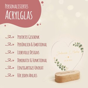 May include: A heart-shaped acrylic glass plaque featuring the printed names Johanna & Markus and the date 28.08.2026, decorated with green eucalyptus leaves. The plaque is mounted on a light-colored wooden base. Text includes Personalisiertes Acrylglas.