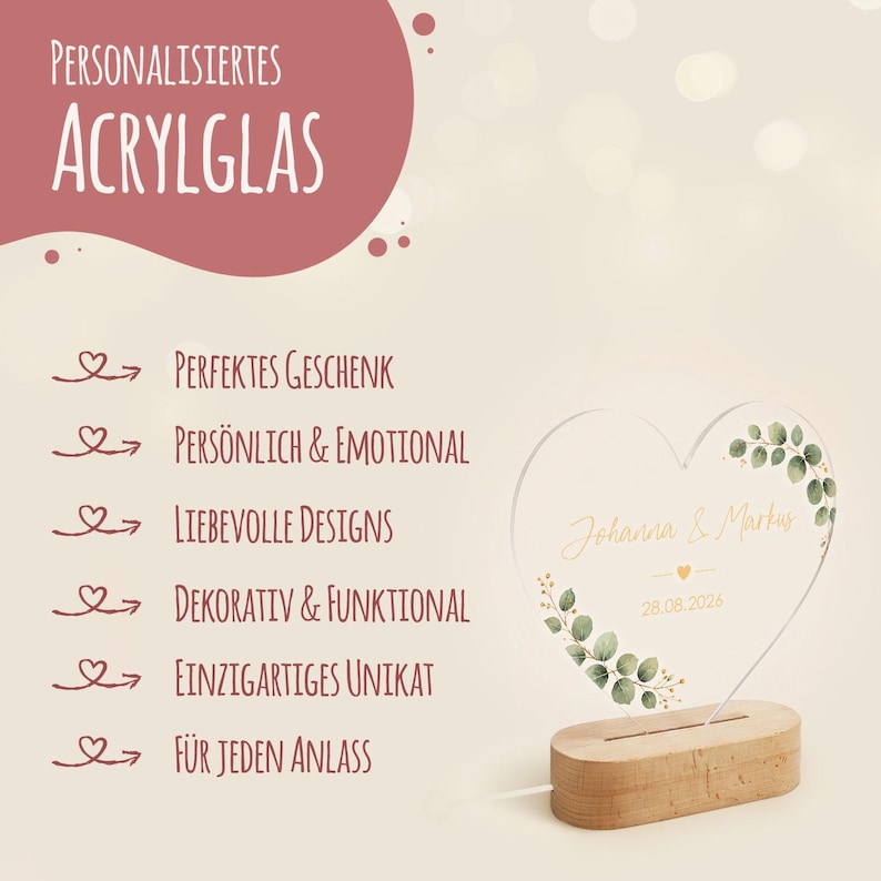 May include: A heart-shaped acrylic glass plaque featuring the printed names Johanna & Markus and the date 28.08.2026, decorated with green eucalyptus leaves. The plaque is mounted on a light-colored wooden base. Text includes Personalisiertes Acrylglas.