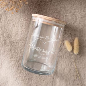 Op de afbeelding: Een heldere glazen pot met een houten deksel. De pot is gegraveerd met de namen "Melanie & Dirk", een hart en de datum "22.12.2025". De pot is cilindrisch en staat op een neutrale ondergrond.