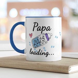 Könnte beinhalten: Weißer Keramikbecher mit blauem Henkel und Rand. Der Becher zeigt den Text "Papa" über einer Babyflaschengrafik mit "2025" darin, einem rosa Herz und den Worten "loading..." darunter. Blaue Fußabdrücke sind um das Design verstreut.