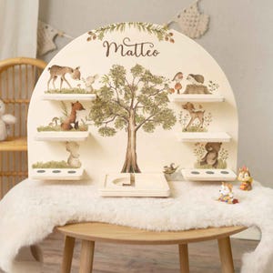 Peut inclure: Étagère en bois sur le thème de la forêt. L'étagère présente une illustration d'arbre, de petites étagères et des figurines d'animaux de la forêt. Le nom "Matteo" est écrit en haut. L'étagère est posée sur une table ronde en bois avec un tapis en fausse fourrure blanche.