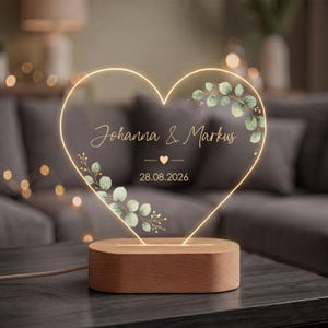 Op de afbeelding: Een hartvormige LED-lamp met een houten voet. Het heldere acrylhart heeft een oplichtende omtrek, groene aquarel eucalyptusbladeren en de tekst "Johanna & Markus" boven een klein hartje en de datum "28.08.2026" in wit cursief schrift.