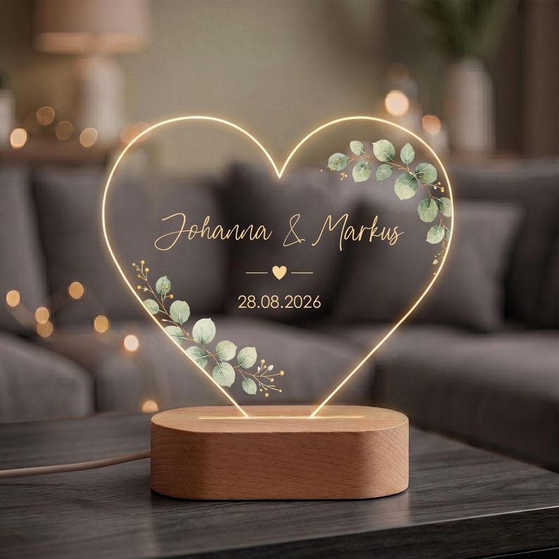 May include: A heart-shaped LED lamp with a wooden base. The clear acrylic heart features a glowing outline, green watercolor eucalyptus leaves, and the text "Johanna & Markus" above a small heart icon and the date "28.08.2026" in white script.