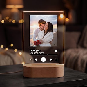 Op de afbeelding: Gepersonaliseerde LED-nachtlamp met foto van een stel, een houten voet en de tekst "Love you, Ich liebe dich!" in het wit. Het licht heeft een ontwerp van een muziekspeler.