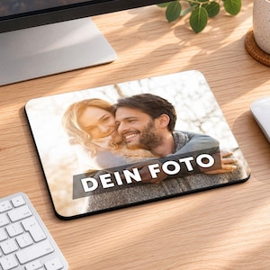 Op de afbeelding: Een rechthoekige muismat met een afgedrukte foto van een koppel dat elkaar buiten omhelst. De afbeelding toont een persoon met lang haar die een persoon met een baard vasthoudt. Een zwarte horizontale balk bevat de witte tekst DEIN FOTO.