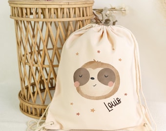 Op de afbeelding: Een beige trekkoordtas met een cartoon luiaard gezicht met gesloten ogen en roze wangen, omringd door kleine sterren. De naam "Louis" staat in sierlijke letters. Een gevlochten rieten mand staat op de achtergrond. De tas is van zacht materiaal.