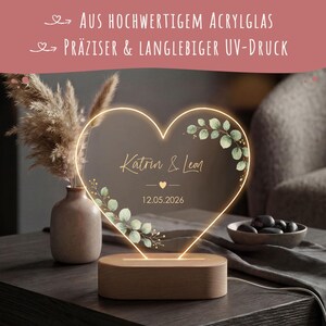 May include: A heart-shaped acrylic light with a wooden base, featuring the printed names Katrin & Leon, the date 12.05.2026, and green leaf illustrations. The top text reads: AUS HOCHWERTIGEM ACRYLGLAS and PR&Auml;ZISER & LANGLEBIGER UV-DRUCK.