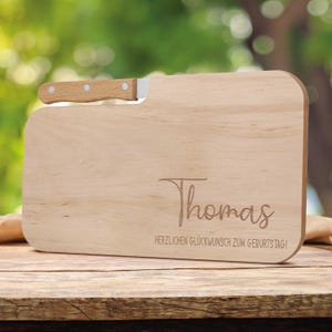 Puede incluir: Una tabla de cortar de madera clara con un cuchillo pequeño adjunto. La tabla tiene esquinas redondeadas y el nombre "Thomas" grabado, junto con el texto "HERZLICHEN GLÜCKWUNSCH ZUM GEBURTSTAG!"