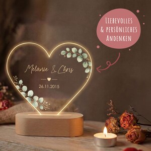 May include: A heart-shaped acrylic light with a warm glow, featuring the names Melanie & Chris, the date 26.11.2015, and green leaf illustrations. The light is mounted on a smooth, oval wooden base, accompanied by a small tea light candle.