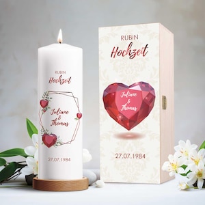 Könnte beinhalten: Eine weiße Säulenkerze mit brennender Flamme, verziert mit einem geometrischen Design mit roten Herzen und den Namen 'Juliane & Thomas'. Eine Holzkiste mit rotem Herzdesign und dem Text 'Rubin Hochzeit' steht neben der Kerze. Das Datum 27.07.1984 ist auf beiden gedruckt.