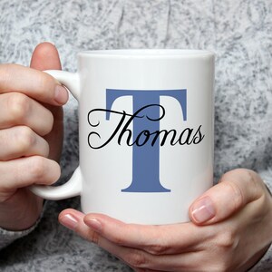 Può includere: Tazza in ceramica bianca con la lettera "T" blu e il nome "Thomas" in corsivo nero. La tazza ha un manico ed è tenuta da una persona che indossa un maglione grigio. Idea regalo personalizzata.