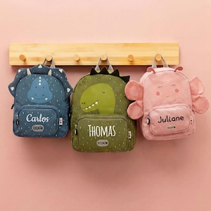 Può includere: Tre zaini per bambini appesi a un attaccapanni di legno. Gli zaini hanno la forma di un dinosauro, un dinosauro e un elefante. Gli zaini a dinosauro sono blu e verdi, e lo zaino a elefante è rosa. Ogni zaino ha un nome.