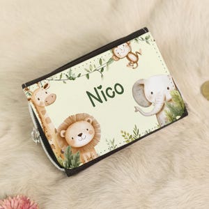 Op de afbeelding: Een kleine portemonnee met ritssluiting, een lichtgele achtergrond en een zwarte rand. De portemonnee heeft illustraties van een giraffe, leeuw, olifant en aap. De naam "Nico" is in het groen gedrukt. Het thema is een safari of jungle.