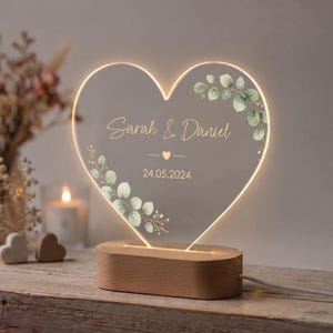 May include: A heart-shaped acrylic lamp with a light-up edge, featuring the printed text Sarah & Daniel and the date 24.05.2024. The design includes watercolor-style green eucalyptus leaves and a small heart icon, set on a light wood base.