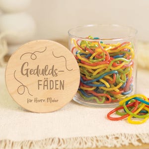 Può includere: Un barattolo di vetro trasparente riempito con fili di plastica colorati, ondulati e simili a spaghetti. Il coperchio rotondo in legno chiaro è inciso con il testo "Gedulds-FÄDEN" e "für Herrn Maier" insieme a linee decorative a spirale.