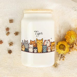 Könnte beinhalten: Ein weißes Keramikglas mit Holzdeckel, verziert mit einer Cartoon-Illustration von Katzen. Das Wort "Tiger" ist kursiv geschrieben. Das Glas ist von katzenförmigen Leckerlis und Trockenblumen umgeben.
