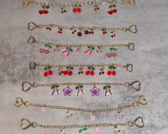 May include: Gold-colored chain bracelets with various charms, including red cherries, pink cherries, and purple flowers. Each bracelet has a heart-shaped clasp. The charms are detailed with green leaves and small crystals.