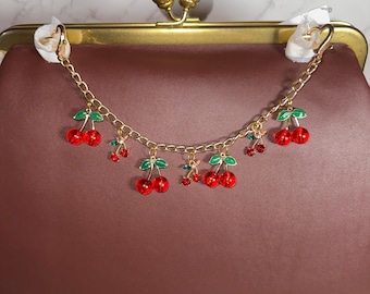 May include: A brown leather clutch bag with a gold clasp and a chain strap adorned with red and green cherry charms.