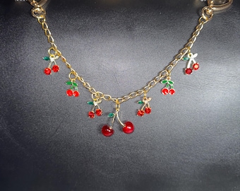 May include: A gold chain with red and green cherry charms hangs from a black leather purse.