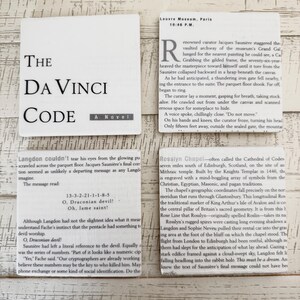 May include: Four white cards with text from the novel "The Da Vinci Code" are arranged on a wooden surface. The cards include excerpts about the Louvre Museum, Rosslyn Chapel, and a coded message.