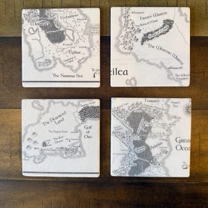 May include: Set of four square coasters featuring detailed black and white fantasy map designs. Each coaster displays a different section of a fictional world, with names of locations and geographical features. The coasters are on a dark wooden surface.