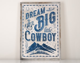 May include: A blue and white framed print with the text "Dream Big Little Cowboy" in a vintage style font. The print features a cowboy riding a horse, a horseshoe, stars, and a mountain range.