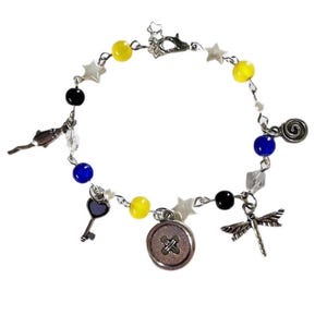 May include: A silver-toned bracelet featuring yellow, blue, and black beads, along with star-shaped charms and decorative pendants. The bracelet includes a key, button, dragonfly, and other charms.