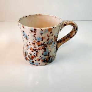 May include: A handmade ceramic mug with a speckled design. The mug features a cream-colored base with blue, brown, and orange speckles. It has a rounded body, a wide opening, and a curved handle. The interior is a light beige color.